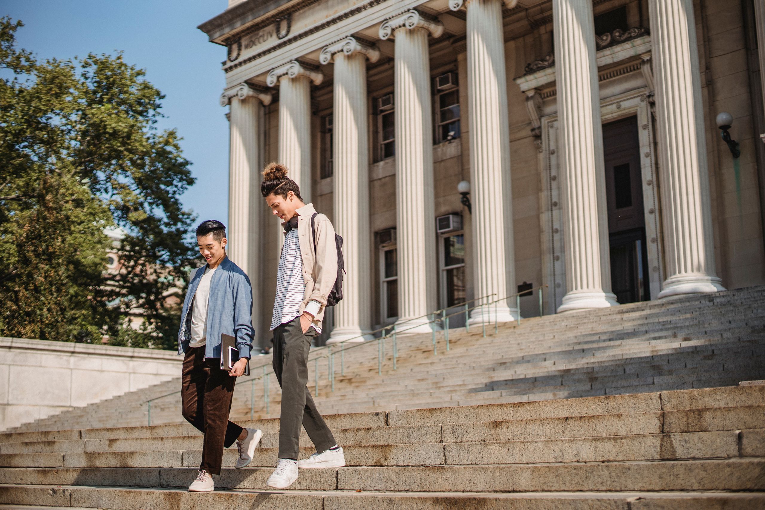 Full body of young multiracial male friends in casual clothes strolling in city on stairs near building with backpack and talking in daytime
